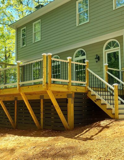 A newly constructed wooden deck with black and white railing, stairs, and lattice skirting attached to a green house with multiple windows.