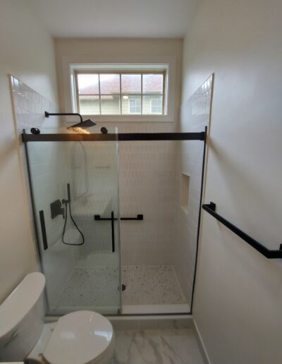 A compact bathroom with a glass-enclosed shower, featuring white tiled walls, a small window, black fixtures, and a toilet in the foreground.