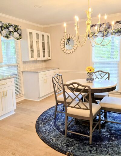 A dining room with a round table, four chairs, and a chandelier. White cabinetry with glass doors and wine cooler on the left. A side table and lamp to the right, with artwork on the wall.