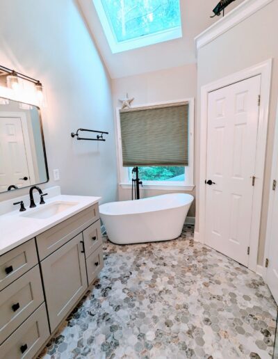 A bright bathroom features a freestanding bathtub, a vanity with a large mirror and a skylight. The space has a hexagonal tiled floor and a window with a blind, providing a view of greenery outside.