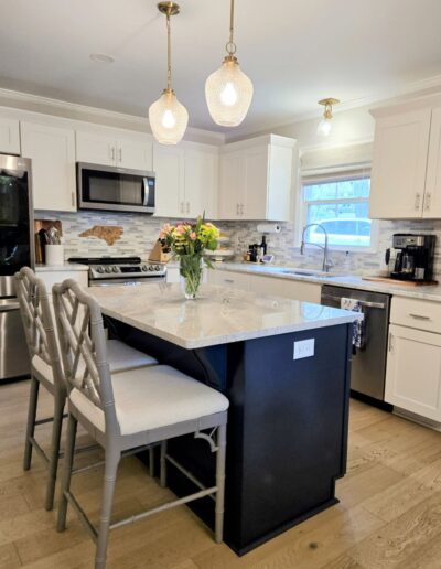 A modern kitchen featuring white cabinets, stainless steel appliances, an island with a marble countertop, two pendant lights, bar stools, a vase with flowers, and wood flooring.