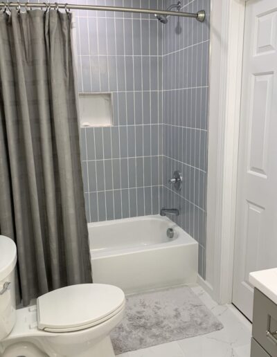 A bathroom with a bathtub and shower, gray tiled walls, gray shower curtain, white toilet, white door, gray rug, and white floor tiles.