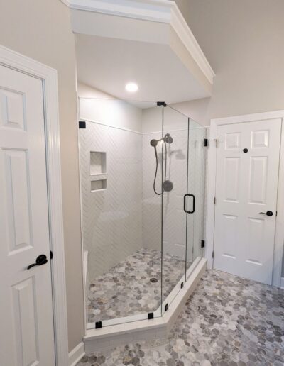 A modern bathroom with a glass-enclosed shower. The shower features gray herringbone tile and a mounted showerhead. The floor is tiled with a hexagonal pattern. White doors are on both sides.