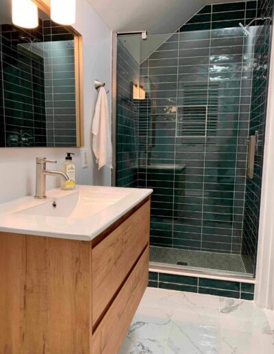 Modern bathroom featuring a wooden vanity with a white countertop, rectangular mirror, and a walk-in shower with dark green tiles. Wall-mounted light fixtures and a towel hanger with a white towel.