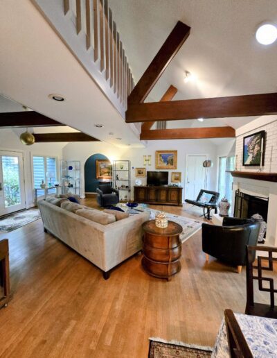 Living room with vaulted ceiling, wooden beams, light-colored sofas, armchairs around a round table, and a fireplace. Decor includes framed pictures and wooden furniture on a wooden floor.